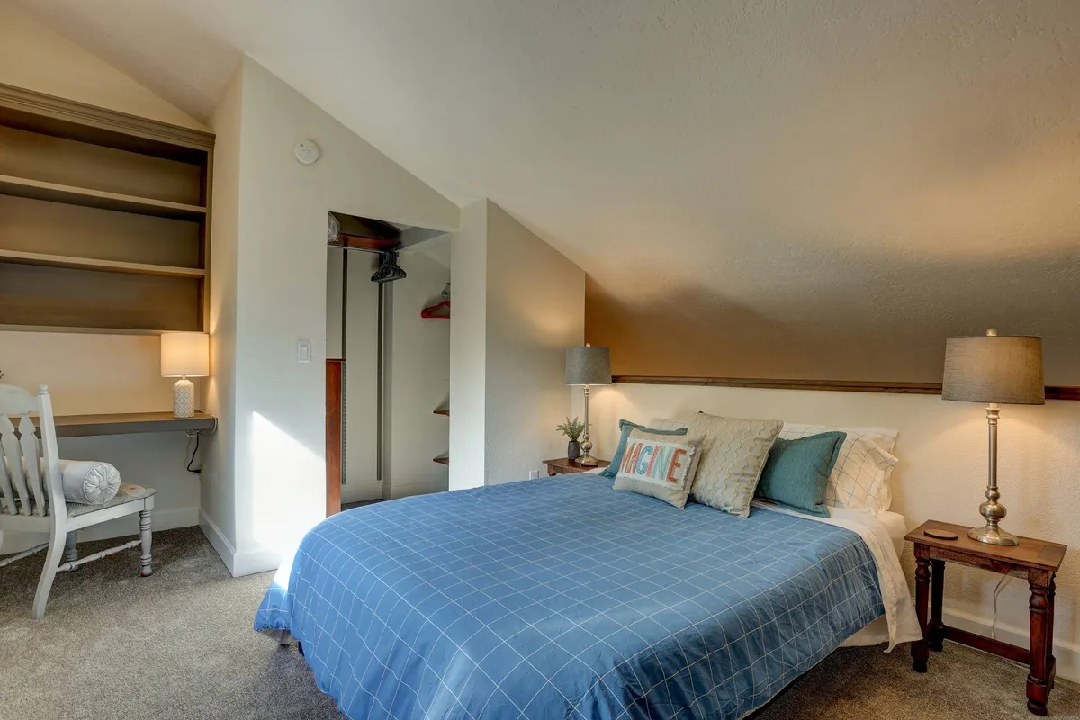 A serene, calming loft space featuring a bed with blue checkered bedding, two bedside lamps, a wooden desk and chair, and shelving. The workspace and natural light evoke a sense of openness and flexibility, fitting for remote work or journaling.
