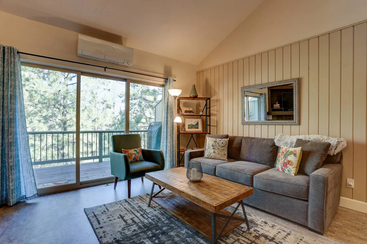 The image showcases a cozy living room featuring a plush sofa, a stylish green chair, a bookshelf, and a coffee table, all bathed in natural light from large windows that frame tranquil views of nature just outside. The setting invites relaxation, reading, and gathering with friends or family while watching sunsets through the windows.