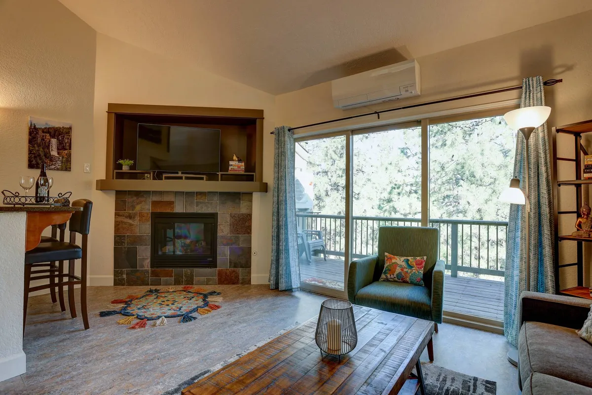 A welcoming living area featuring a prominent fireplace, comfortable seating, large windows that fill the room with natural light, and a nearby dining area. The mood is warm and inviting, ideal for both relaxation and socializing.
