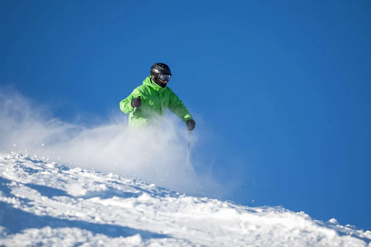 A skier in a bright green jacket glides down a snow-covered slope beneath a vibrant blue sky. In the distance, pine trees and mountains frame the exhilarating winter scene.