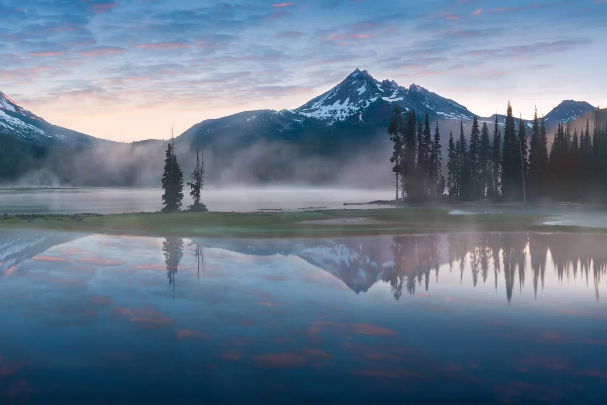 A tranquil alpine lake sits at the foot of misty mountains, with pine trees framing the view. Early morning fog drifts across the water, creating a peaceful and awe-inspiring landscape perfect for exploring, hiking, or simply enjoying the scenery.