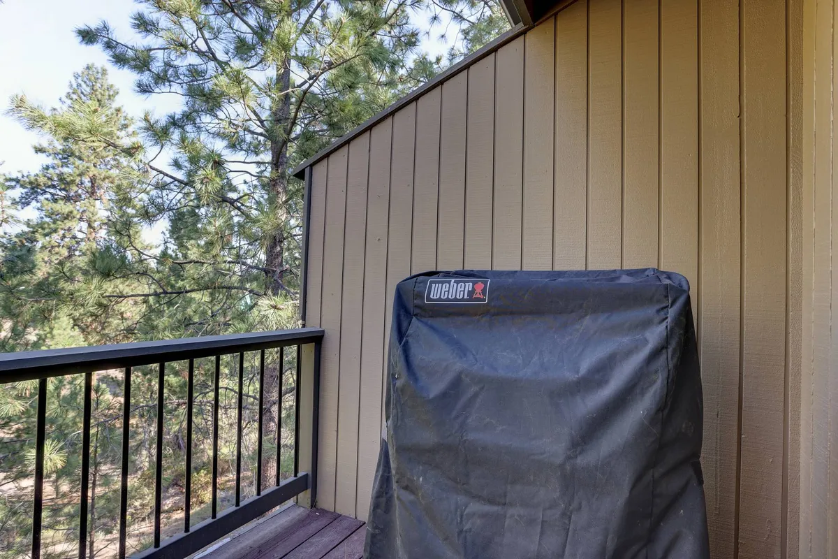 This image shows a wooden deck surrounded by lush pine trees, featuring a covered grill and wooden railings. The casual setup is ideal for barbecuing or enjoying outdoor meals with a backdrop of natural beauty.