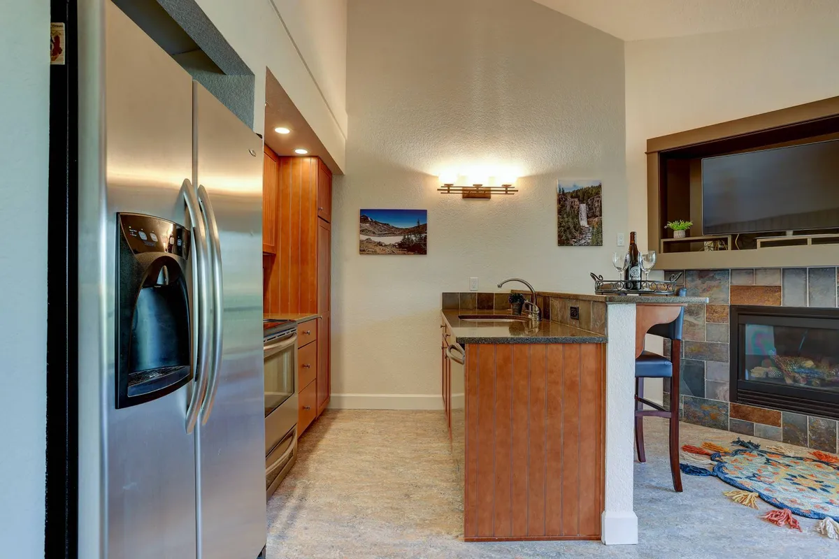 A spacious, sunlit great room seamlessly combining an open-concept living area and kitchen. The image captures stainless steel appliances, a central kitchen island, and a cozy fireplace, with the setting arranged for cooking, entertaining, and family gatherings.
