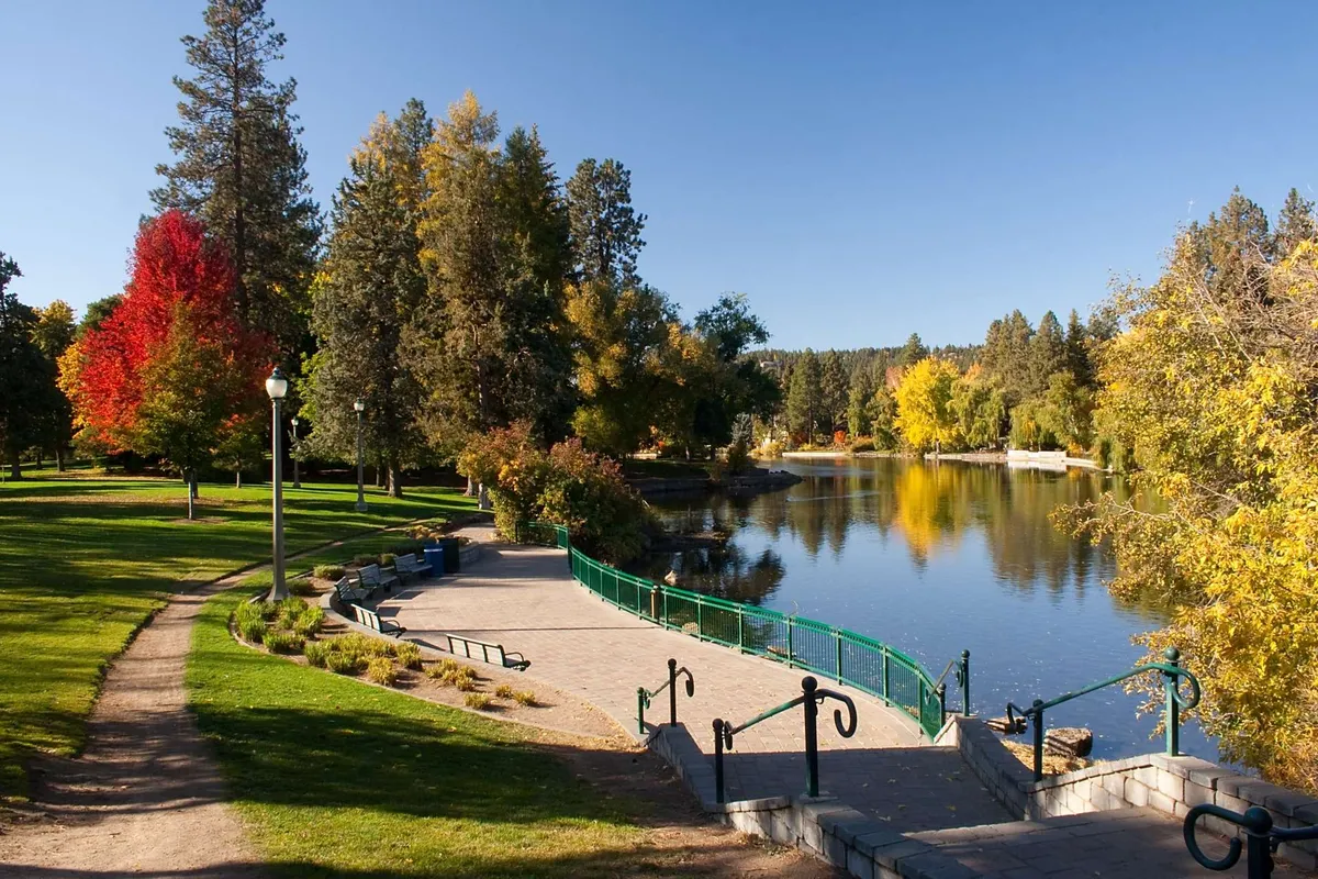 A serene lake with colorful trees, a walking path, and park benches creates an inviting backdrop for relaxation, walks, and picnics. The image radiates tranquility and the appeal of year-round outdoor amenities.