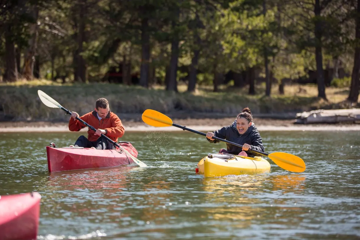 Two kayakers paddle through calm waters, surrounded by lush greenery and natural riverbanks. The brilliant colors of the kayaks add energy to the serene, sunlit setting, capturing the joy and adventure of water activities on the Deschutes River.