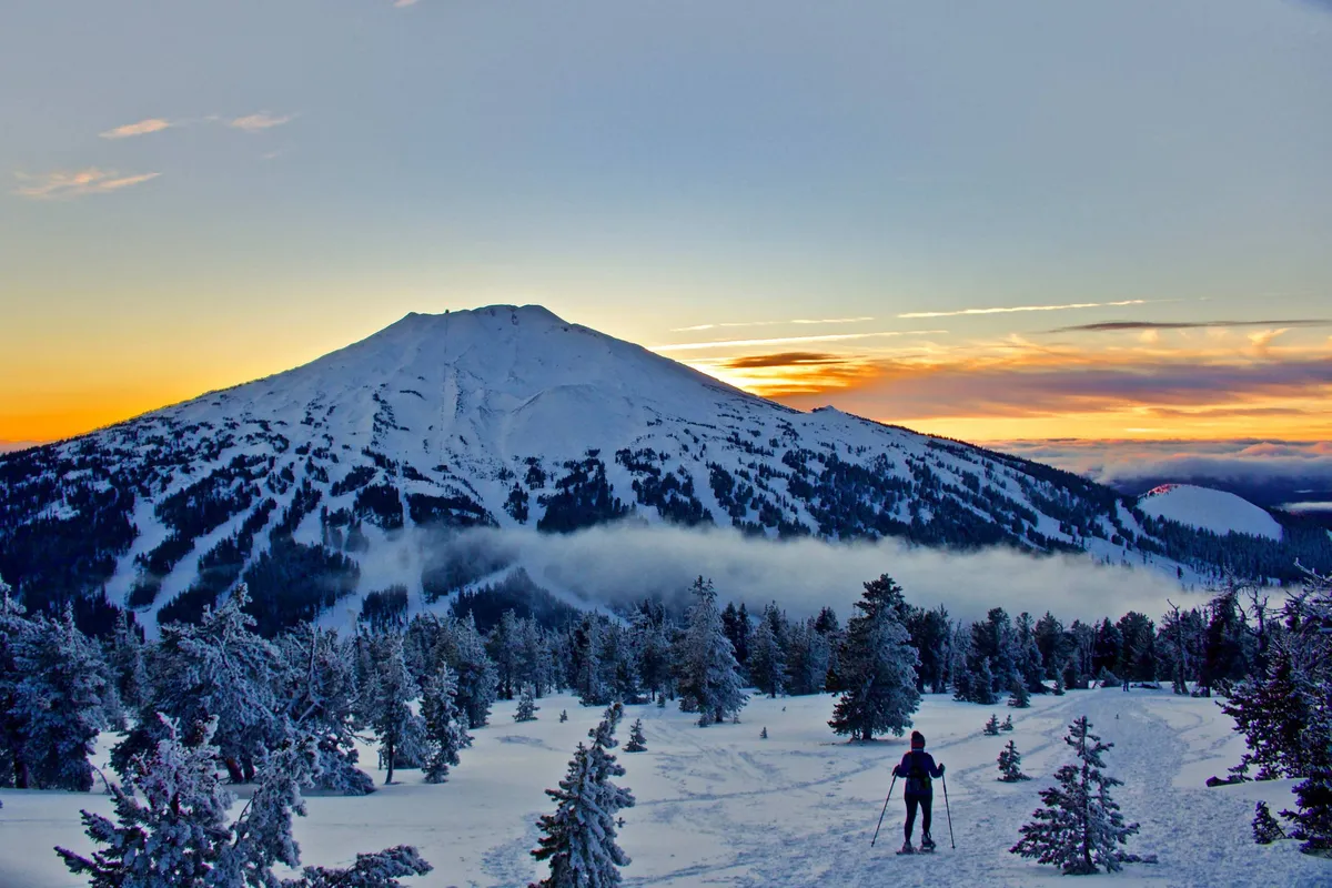 A snow-covered mountain dominates the scene, with a skier making fresh tracks down the slope under a glowing sunset sky. The expansive winter landscape captures the thrill and beauty of outdoor adventure in snowy conditions.