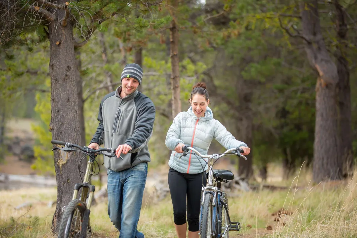 Two cyclists walk their bikes along a path lined with tall trees and vibrant greenery, suggesting active outdoor fun and family-friendly recreation just steps from the resort.